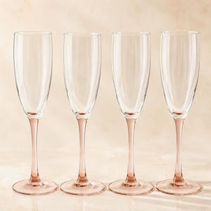 Peut inclure: Un ensemble de quatre flûtes à champagne en verre transparent, chacune dotée d'une tige fine et d'une base circulaire teintées d'un rose pâle délicat. Les coupes sont hautes et étroites, conçues pour les boissons pétillantes.