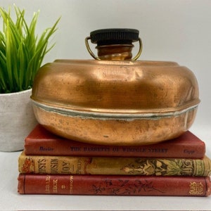 May include: A vintage copper water bottle with a black plastic lid and a brass spout. The bottle is sitting on a stack of old books.