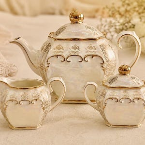 May include: An ivory porcelain tea set with gold detailing. The set comprises a teapot, a creamer jug, and a lidded sugar bowl. The pieces have a pearlescent finish and are decorated with gold patterns, ideal for afternoon tea.