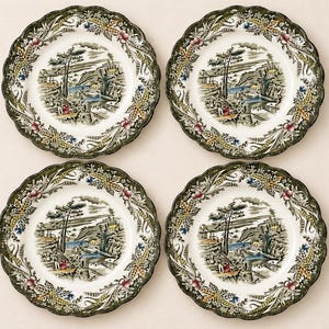 May include: A set of four round ceramic plates with scalloped edges. Each plate features a central landscape illustration in black, white, and blue, surrounded by a wide border decorated with colorful wheat, leaves, and berries in green and red.