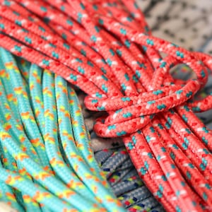 May include: A collection of braided cords featuring various colors and patterns. The foreground displays a bright coral cord with small teal and white diamond accents, while a turquoise cord with yellow and orange patterns sits to the left.