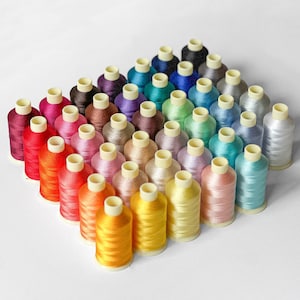 May include: A collection of forty-two spools of sewing thread arranged in a grid pattern. The spools feature a wide spectrum of colors including shades of red, orange, yellow, green, blue, purple, pink, brown, gray, and white with white caps.