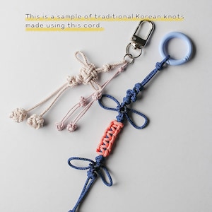 May include: Two decorative cord ornaments featuring traditional Korean knots. One is a light beige cord with a silver metal lobster clasp. The other is a blue cord with a light blue plastic ring and a central section of coral-colored woven cord.
