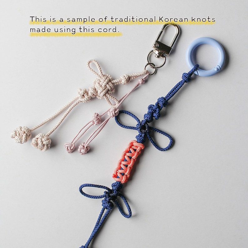 May include: Two decorative cord ornaments featuring traditional Korean knots. One is a light beige cord with a silver metal lobster clasp. The other is a blue cord with a light blue plastic ring and a central section of coral-colored woven cord.