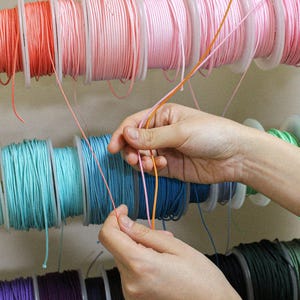 May include: Multiple spools of thin, smooth cord in a variety of colors including peach, pink, turquoise, blue, and green are arranged on horizontal racks. Two strands of cord, one peach and one pink, are being held and pulled forward.