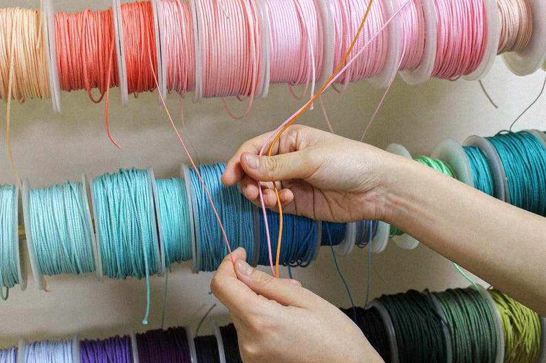May include: Multiple spools of thin, smooth cord in a variety of colors including peach, pink, turquoise, blue, and green are arranged on horizontal racks. Two strands of cord, one peach and one pink, are being held and pulled forward.
