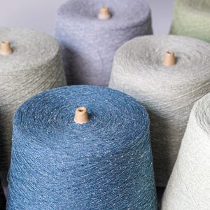 May include: A collection of yarn cones in various colors including deep blue, charcoal gray, light gray, sage green, and lavender. Each conical spool is tightly wound with textured, speckled yarn and features a small wooden center peg at the top.