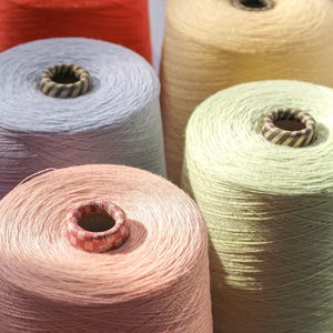 May include: Five cylindrical spools of yarn arranged in a cluster. The colors include a vibrant orange, a soft yellow, a light periwinkle blue, a pale mint green, and a muted peach. Each spool features a central cardboard core with a patterned rim.