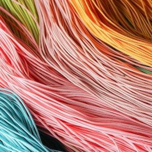 May include: A collection of thin, smooth, multi-colored nylon cords arranged in parallel lines. The strands feature vibrant shades of turquoise, light pink, soft peach, and bright orange, creating a textured, layered appearance of fine, flexible fibers.
