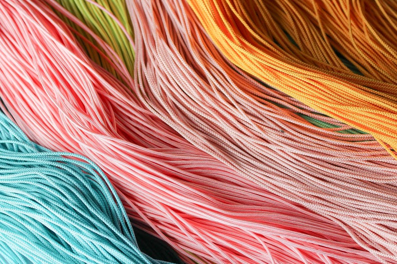 May include: A collection of thin, smooth, multi-colored nylon cords arranged in parallel lines. The strands feature vibrant shades of turquoise, light pink, soft peach, and bright orange, creating a textured, layered appearance of fine, flexible fibers.