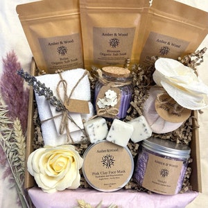 May include: A gift box filled with bath and body products from Amber & Wood. The box includes a white towel, a jar of purple bath salts, a jar of pink clay face mask, a jar of violet whipped cream soap, and two white soap bars. The box is decorated with lavender sprigs and a white paper flower.