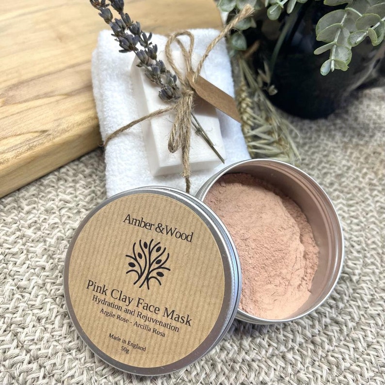 May include: A brown paper label with the text "Amber & Wood" and "Pink Clay Face Mask" with a small tree illustration. The label is attached to a silver tin containing a pink clay face mask. The tin is 50g and made in England.