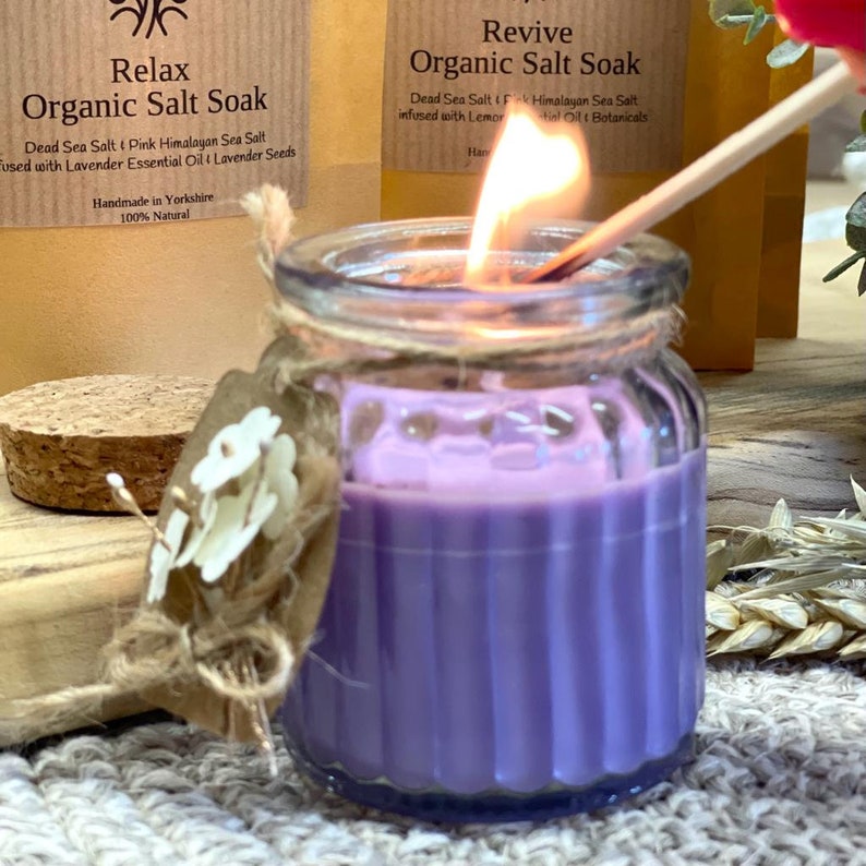 May include: A purple candle in a clear glass jar with a cork lid. The candle is lit and the flame is visible. The jar has a label that says "Relax Organic Salt Soak" and "Dead Sea Salt & Pink Himalayan Sea Salt used with Lavender Essential Oil & Lavender Seeds". The label also says "Handmade in Yorkshire 100% Natural".