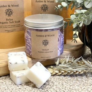 May include: A glass jar filled with purple bath salts labeled "Amber & Wood Violet Whipped Cream Soap Made in Yorkshire". Three white soap bars with dried flowers are in the foreground.