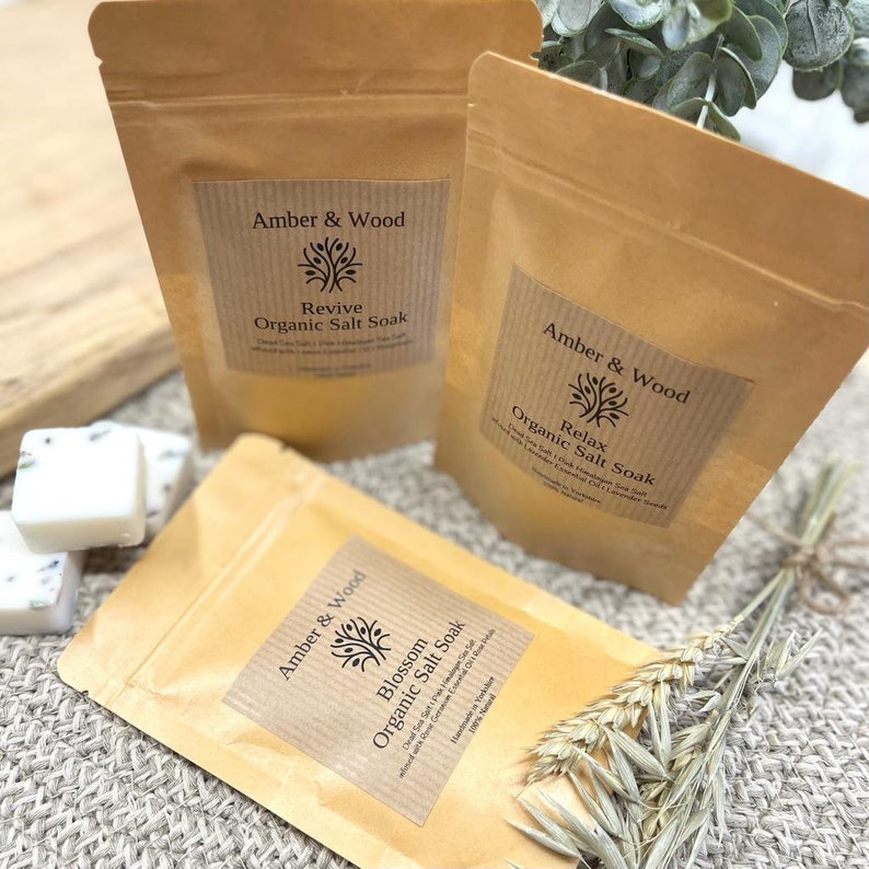May include: Three brown paper bags with labels that read "Amber & Wood" and different scents: "Revive", "Relax", and "Blossom". Each bag contains organic salt soak.
