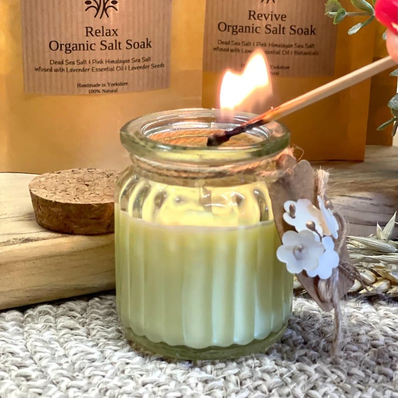 May include: A clear glass jar candle with a yellow wax interior and a cork lid. The candle is lit with a match and has a brown twine tied around the jar with a small white flower decoration.