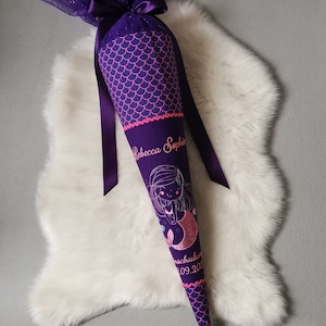 Könnte beinhalten: Lila-rosa Schulschultüte mit Meerjungfrauen-Motiv. Auf der Schultüte sind der Name "Rebecca Sophie" und das Datum "09.202..." gestickt. Die Schultüte ist mit einer lila Schleife aus Tüll und einem lila Band verziert.