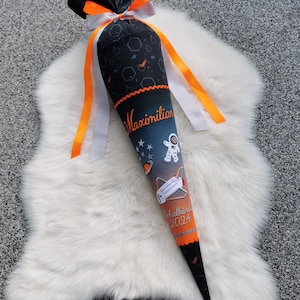 Könnte beinhalten: Eine schwarz-orange Schultüte in Kegelform mit Weltraum-Motiv. Die Tüte zeigt einen Astronauten, eine Rakete und den Namen "Maximilian". Sie ist mit orangefarbenen und weißen Bändern sowie dem Text "Schulkind 2024" verziert.