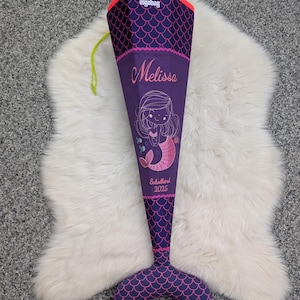 Könnte beinhalten: Schultüte in Lila mit Meerjungfrauen-Motiv. Sie zeigt eine Meerjungfrauen-Illustration, den Namen "Melissa" und den Text "Schulkind 2025". Der obere Teil hat eine rosa Bordüre und ein Fischschuppenmuster.