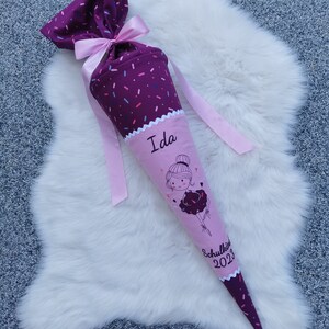 Könnte beinhalten: Eine lila-rosa Schultüte mit einem Ballerina-Motiv und dem Namen "Ida". Sie hat eine rosa Schleife und ein Band, mit dem Text "Schulkind 2023". Die Tüte liegt auf einer weißen, flauschigen Oberfläche.