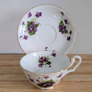 May include: White teacup and saucer set with a floral violet design. The teacup has a curved handle and a gold rim. The saucer has a matching violet pattern and a gold rim. The set is on a wooden surface.
