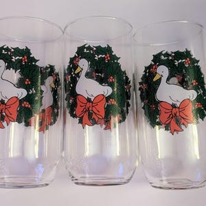 May include: Three clear glass tumblers with a festive design. Each glass features a white duck with a yellow beak, surrounded by a green wreath with red berries and a large red bow. The glasses are ideal for holiday drinks.