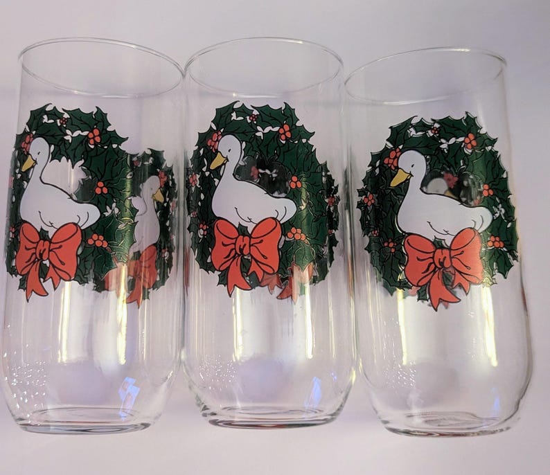 May include: Three clear glass tumblers with a festive design. Each glass features a white duck with a yellow beak, surrounded by a green wreath with red berries and a large red bow. The glasses are ideal for holiday drinks.