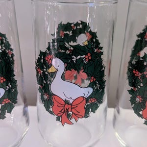 May include: Clear glass tumblers with a festive design. Each glass features a white duck inside a green holly wreath with red berries and a red bow. The glasses are ideal for holiday beverages.