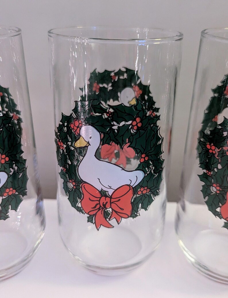 May include: Clear glass tumblers with a festive design. Each glass features a white duck inside a green holly wreath with red berries and a red bow. The glasses are ideal for holiday beverages.