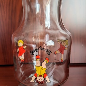 May include: A clear glass pitcher with a yellow lid and a Cabbage Patch Kids logo. The pitcher features three Cabbage Patch Kids characters, one of which is upside down.