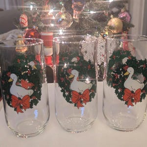 May include: Three clear glass tumblers with a Christmas design. Each glass features a white duck with a yellow beak, a red bow, and a green wreath with red berries. The glasses are in front of a Christmas tree.