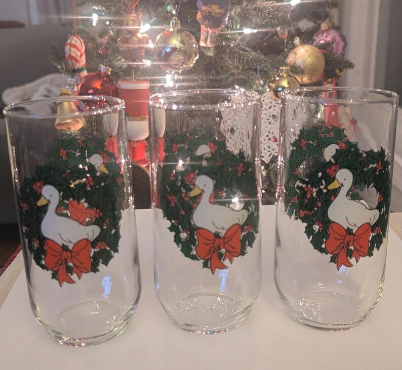 May include: Three clear glass tumblers with a Christmas design. Each glass features a white duck with a yellow beak, a red bow, and a green wreath with red berries. The glasses are in front of a Christmas tree.