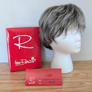 Könnte beinhalten: Eine kurze, gestufte Perücke in Grau- und Hellbrauntönen wird auf einem weißen Styroporkopf präsentiert. Eine rote Schachtel mit dem Markennamen "Jon Renau" und eine Produktinformationskarte befinden sich neben der Perücke. Die Karte zeigt den Stil und die Farbe der Perücke.