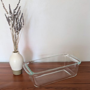 Peut inclure: Un moule à pain rectangulaire en verre transparent avec des poignées intégrées aux extrémités courtes, placé à côté d'un petit vase en céramique crème moucheté contenant un bouquet de tiges de lavande séchées.