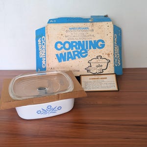 May include: A white Corning Ware covered saucepan with a blue floral pattern, featuring a clear glass lid and cardboard packaging. The box displays the text Corning Ware, Makes Life Easier, Cook on the Range Top, in the Oven, or Microwave Oven.