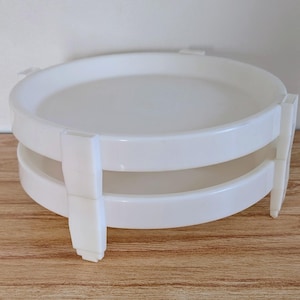 Könnte beinhalten: Ein weißes, zweistufiges Serviertablett mit kreisförmigem Design. Das Tablett hat eine glatte, glänzende Oberfläche und wird von vier rechteckigen Beinen getragen. Es besteht aus strapazierfähigem Kunststoff und eignet sich zum Servieren von Speisen oder Präsentieren von Gegenständen.