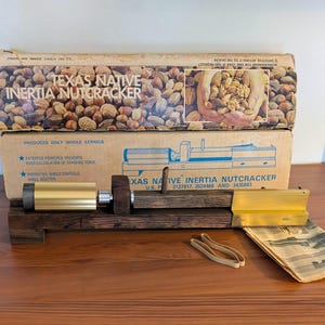 May include: A Texas Native Inertia Nutcracker featuring a dark wood base, a gold-coloured metal cylinder, and a sliding mechanism. The device is presented with its original cardboard packaging, which displays product text and imagery, alongside two rubber bands.