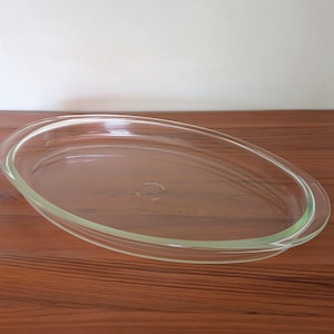 May include: A clear, oval-shaped glass baking dish with integrated side handles. The dish features a smooth, transparent surface and a subtle embossed circular logo mark centered on the interior base, designed for oven use and food preparation.