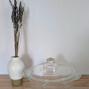 May include: A clear glass casserole dish with a lid, and a small white vase with dried lavender. The casserole dish is on a wooden surface. The vase is white with a gray base.