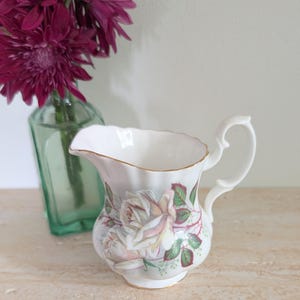 以下が含まれることがあります： 白い磁器のクリーマー。バラの花柄と金の縁取りが施されています。クリーマーは湾曲したハンドルと注ぎ口が付いています。濃い紫色の花のブーケが、緑色のガラス瓶に入って背景にあります。