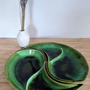 May include: A vintage green ceramic serving dish with three sections. The dish has a glossy finish with dark green and black tones. A small white vase with dried lavender is in the background.