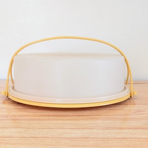 Puede incluir: Un portatartas redondo amarillo y blanco con asa. El soporte tiene una base amarilla y una tapa transparente esmerilada. El asa también es amarilla. El soporte está sobre una superficie de madera. Aproximadamente 25 cm de diámetro.