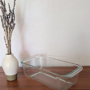 Könnte beinhalten: Eine rechteckige Kastenform aus klarem Glas mit integrierten Griffen an den kurzen Seiten, platziert neben einer kleinen, gesprenkelten cremefarbenen und beigen Keramikvase mit einem Bündel getrockneter Lavendelstiele mit grauvioletten Blüten.