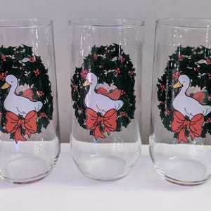 May include: Set of three clear glass tumblers. Each tumbler features a white duck with a yellow beak, a red bow, and a green wreath design. The tumblers are ideal for serving beverages.