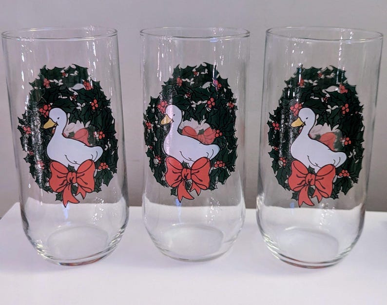 May include: Set of three clear glass tumblers. Each tumbler features a white duck with a yellow beak, a red bow, and a green wreath design. The tumblers are ideal for serving beverages.
