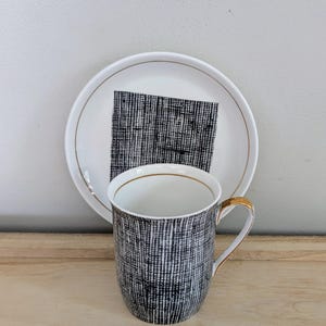 Puede incluir: Un juego de taza y platillo de cerámica blanca. La taza y el platillo tienen un patrón texturizado en blanco y negro. La taza tiene un asa dorada y ribete dorado. El platillo tiene un borde dorado y un diseño cuadrado en blanco y negro.