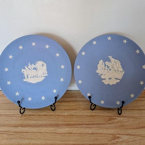 May include: Two blue decorative plates with white star accents and historical scenes. One plate reads "America's Heritage" with a covered wagon illustration. The other plate reads "American Independence 1776" with a battle scene.