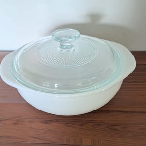 May include: A round white casserole dish with integrated side handles and a clear glass lid. The lid features a central knob handle and a textured radial pattern radiating from the center, providing a clear view into the white bowl base.