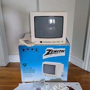 May include: A vintage Zenith 9-inch (approx. 23 cm) colour receiver/monitor in Arctic White. The TV is on its original box, with a remote, antenna, and accessories. The box features the Zenith logo and product details. The TV has a small screen and a classic design.