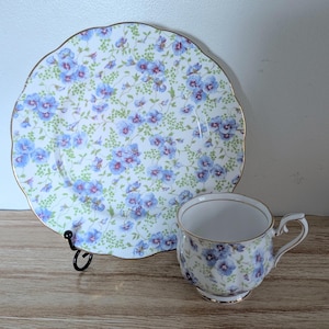 May include: A porcelain tea cup and matching saucer featuring a white base with a repeating pattern of small blue flowers and green foliage. Both pieces have scalloped edges and are finished with a thin gold trim along the rims and handle.